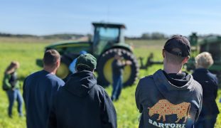 A speaker presents in front of a green tractor as a group listens in a grassy field — highlighting hands-on agricultural education during a farm demonstration.