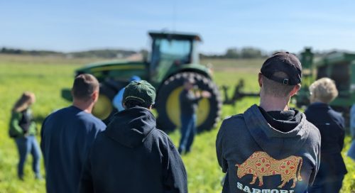 A speaker presents in front of a green tractor as a group listens in a grassy field — highlighting hands-on agricultural education during a farm demonstration.