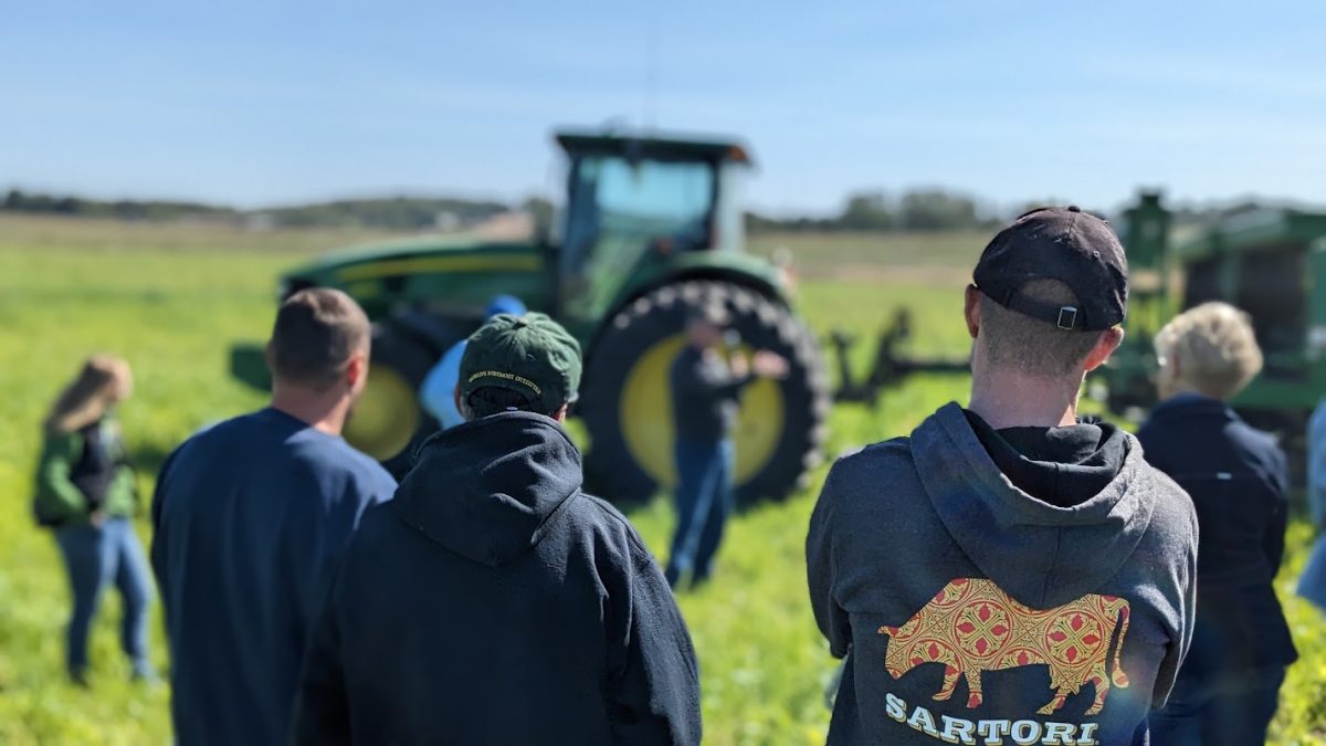 A speaker presents in front of a green tractor as a group listens in a grassy field — highlighting hands-on agricultural education during a farm demonstration.